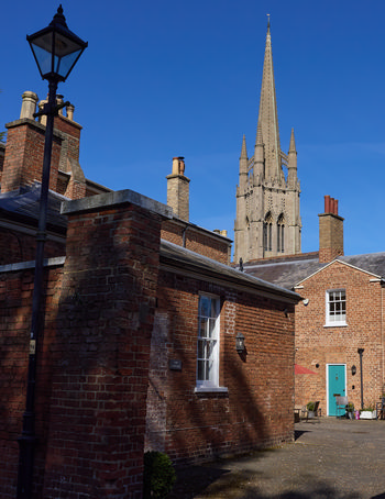 This urban architecture photograph taken in the afternoon during spring in Louth, Lincolnshire, features the brick chimneys of buildings near St James' Church as its main subject. The tall stone spire of St James' Church, a prominent landmark in Louth and Lincolnshire, rises in the background under a clear blue sky. The image highlights the architectural details typical of the area, including red brickwork and a traditional streetlamp, with the church contributing to the historic atmosphere of the scene.